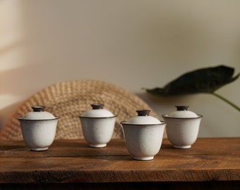 May include: Four white ceramic tea cups with lids. Each cup has a speckled texture and a dark brown rim and lid. The cups are arranged on a wooden surface, with a woven mat and a large leaf in the background.