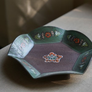 May include: A hexagonal ceramic dish with a green and brown glaze. The dish has a floral design with orange and white accents.