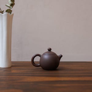 May include: A small, brown teapot with a rounded body and a small knob on the lid. The teapot is sitting on a dark brown wooden surface.