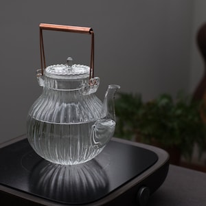 Pode incluir: Uma chaleira de vidro transparente com uma alça de cobre e um design canelado. A chaleira está cheia de água e está sobre uma placa de fogão preta. O fundo é cinza desfocado com uma guitarra e uma planta.