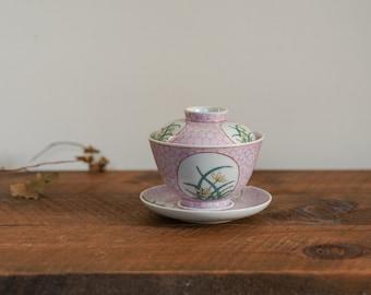 May include: A pink and white porcelain teacup and saucer set with a floral design. The cup has a lid and the saucer has a scalloped edge.