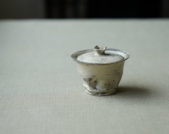 May include: A small, white ceramic teapot with a lid. The pot has a rough, textured surface and a slightly uneven shape. The lid has a small, pointed knob on top.