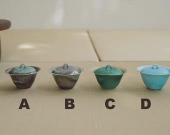 May include: Four small, lidded ceramic bowls in shades of green, brown, and blue are arranged in a row. Each bowl has a matching lid with a small loop handle. The bowls are labelled A, B, C, and D.