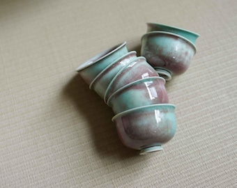 May include: A stack of six ceramic tea cups with a speckled teal and maroon glaze. The cups are stacked on top of each other, with a few more cups to the side. The cups have a small foot and a slightly flared rim.
