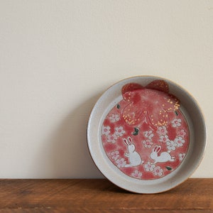 May include: A ceramic plate with a red floral design and two white rabbits. The plate has a beige rim and a gold-colored edge. The plate is on a wooden surface.