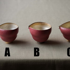 May include: Three handmade ceramic bowls with a distinctive spout design. The bowls are a deep red colour with a contrasting light interior and a small, rounded base. The letters A, B, and C are printed below each bowl.