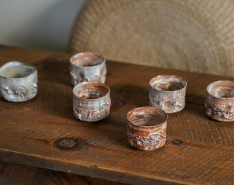 May include: Six small ceramic cups with a white glaze and brown accents. The cups have a textured surface and are arranged on a wooden table.