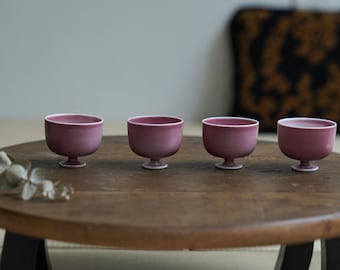 May include: Four small, maroon-colored ceramic cups with white rims and pedestal bases are arranged on a round, wooden table. The cups are evenly spaced, showcasing a minimalist design. The table has black legs, and a decorative pillow is in the background.