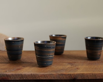 May include: Four dark blue ceramic cups with a brown and gold speckled glaze. The cups are arranged in a row on a wooden surface.