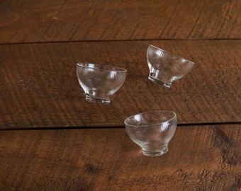 May include: Three clear glass bowls with a ribbed texture sit on a wooden surface.