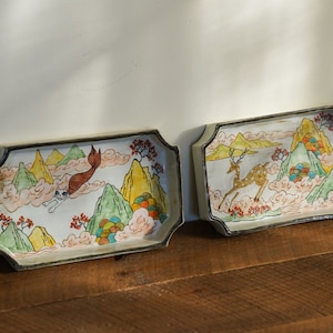 Puede incluir: Dos bandejas rectangulares de cerámica con fondo blanco y diseños pintados de colores. Una bandeja presenta una sirena nadando en las nubes sobre una cordillera, mientras que la otra bandeja presenta un ciervo corriendo a través de una cordillera similar.
