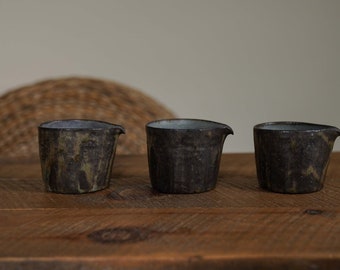 May include: Three small ceramic pitchers with a dark brown glaze and a light gray interior. The pitchers have a slightly flared lip and a rounded base.