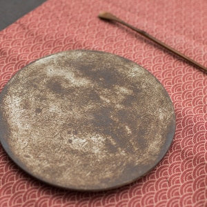 May include: A rustic brown ceramic plate with a white glaze, sitting on a red tablecloth with a white wave pattern.