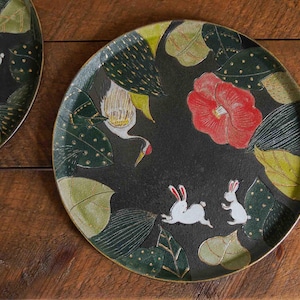 Könnte beinhalten: Ein schwarzer runder Teller mit einem Design aus einem weißen Kranich, weißen Kaninchen und einer roten Blume. Der Teller hat ein Muster aus grünen und goldenen Blättern.