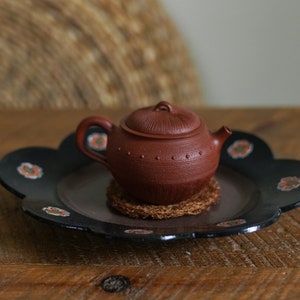 May include: A small, red clay teapot with a lid sits on a black, flower-shaped plate with a woven coaster underneath. The teapot has a textured surface and a small spout.