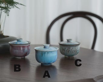 May include: Three ceramic tea cups with lids in shades of blue, purple, and green sit on a dark wooden table. The cups have a rounded shape and a glossy finish. A small plant is visible in the background.