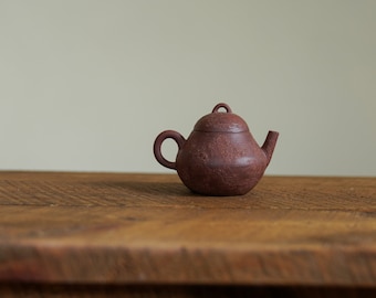 May include: A small, brown, ceramic teapot with a lid. The teapot is sitting on a wooden table.
