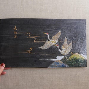 Puede incluir: Panel de madera negro con un diseño pintado en oro y blanco de dos grullas volando sobre una cordillera. El texto "松鶴延年" está escrito en oro en el panel.