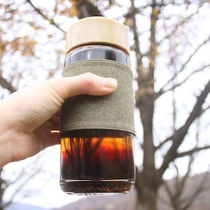 May include: A glass bottle with a brown liquid inside, a beige lid, and a green fabric sleeve. The bottle is being held by a hand.