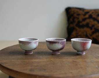 May include: Three small, handcrafted ceramic bowls with a footed base. The bowls have a two-tone color scheme, with a light blue base and a dark purple rim. The bowls are arranged on a round wooden table.