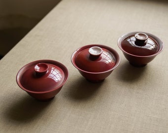 Puede incluir: Tres tazas de té rojas con tapas. Las tapas tienen un interior blanco y una pequeña perilla en la parte superior. Las tazas de té están dispuestas en una fila sobre una superficie marrón claro.