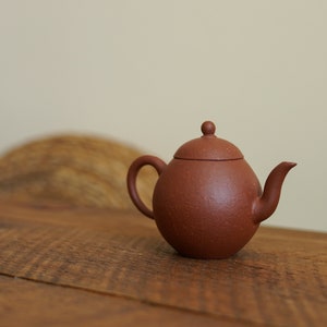 May include: A small, brown clay teapot with a rounded body and a lid. The teapot is sitting on a wooden table.