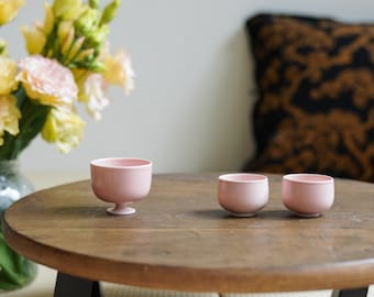 May include: Three small, light pink ceramic cups on a round wooden table. One cup has a small pedestal base. A vase of yellow and pink flowers is in the background.