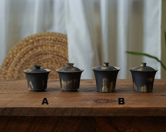 May include: Four black ceramic teacups with gold accents and lids. The teacups are arranged in a row on a wooden surface. The teacups are labelled A and B.