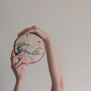 May include: A round, beige tea cake with a printed design of a tree and clouds. The text "2019" and "Sunlit Forest" are printed on the cake.