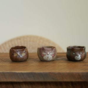 May include: Three small ceramic cups with cat faces painted on them. The cups are brown with white and pink details. They are arranged in a row on a wooden surface.