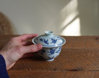 Könnte beinhalten: Eine weiße Porzellan-Teetasse mit Deckel, verziert mit einem blauen und weißen Schmetterlingsmuster. Die Teetasse steht auf einem Holztisch.