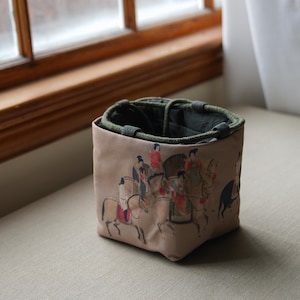 May include: A small, beige fabric bucket with a green drawstring top. The bucket is decorated with a colorful print of people riding horses.
