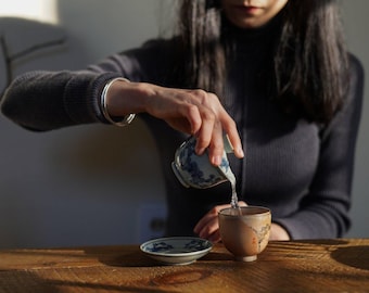 May include: A person pouring tea from a blue and white ceramic teapot into a brown ceramic teacup. The teacup is sitting on a blue and white ceramic saucer. The person is wearing a dark grey sweater.