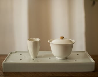 May include: A white ceramic tea set with a lidded teapot and a small cup, both with a fluted design, sitting on a white ceramic tray with holes.