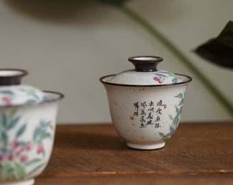 May include: Two white ceramic teacups with lids. The teacups have a floral design with green leaves and pink flowers. One teacup has Chinese calligraphy on the side. The teacups are sitting on a wooden surface.