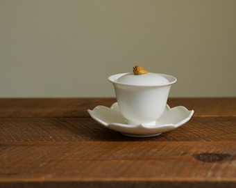 May include: A white ceramic teacup with a lid and a small gold knob on top. The teacup is sitting on a white saucer with a scalloped edge. The teacup and saucer are on a wooden table.