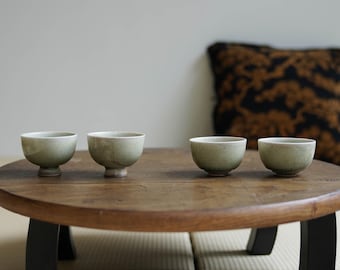 May include: Four small, pale green ceramic tea bowls sit on a round, wooden table with black legs. The bowls have a simple, rounded shape and a slightly textured surface. A decorative pillow with a black and brown pattern is in the background.