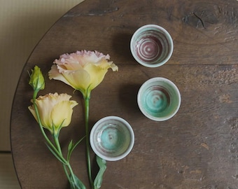 May include: Three small, hand-painted ceramic bowls with a spiral design in shades of teal, brown, and white are arranged on a dark wooden surface. Two yellow and pink flowers and a bud are also on the surface, creating a natural, artistic composition.