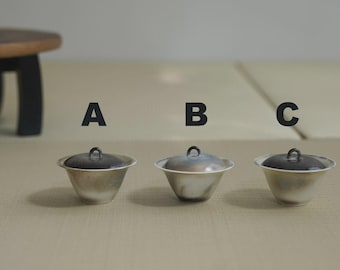 May include: Three small, silver-toned bowls with dark lids are arranged in a row. Each bowl has a rounded shape and a small handle on the lid. The bowls are labeled A, B, and C. A wooden stool is in the background.