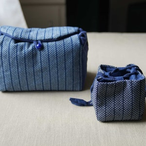 Op de afbeelding: Twee blauwe en witte stoffen tassen met een patroon. De grotere tas heeft een blauwe knoopsluiting. De kleinere tas is gevouwen en heeft een trekkoord.