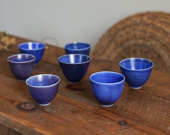 May include: A set of seven small blue ceramic teacups arranged on a wooden table. The cups are all the same size and shape, and they have a slightly textured surface.