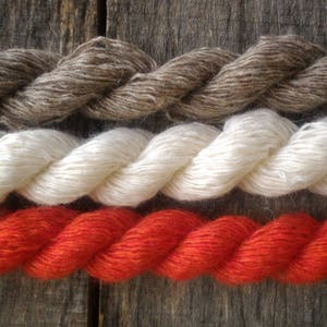 May include: Three strands of yarn in different colors: brown, white, and orange. The yarn is twisted and laid out on a wooden surface.