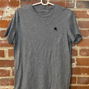 May include: A heather gray crew neck t-shirt with short sleeves. The shirt has a small black embroidered logo on the left chest and the brand name "EXPRESS" printed on the collar.