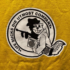 May include: A white circle with black text that reads "Life Sucks Die Utmost Company." Inside the circle is a black and white cartoon of a squirrel wearing a hat and holding a boombox.