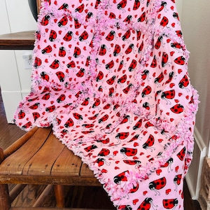 May include: A pink blanket with a ladybug and heart pattern. The blanket has a pink fringe border and is draped over a wooden chair. The ladybugs are red with black spots and antennae.