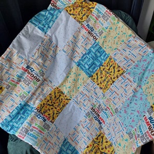 Puede incluir: Una colcha de retazos en colores azul, blanco y amarillo. La colcha est&aacute; hecha de diferentes telas con varios patrones, incluyendo un patr&oacute;n repetido de las palabras "Buzz", "Woody", "Rex" y "Ducky".