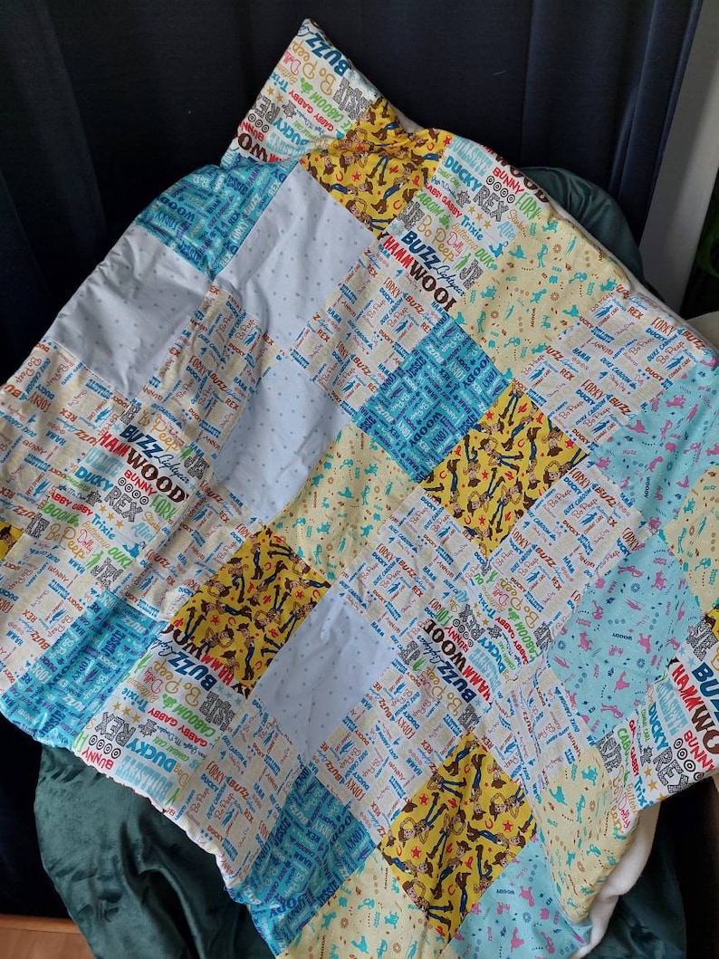 Puede incluir: Una colcha de retazos en colores azul, blanco y amarillo. La colcha est&aacute; hecha de diferentes telas con varios patrones, incluyendo un patr&oacute;n repetido de las palabras "Buzz", "Woody", "Rex" y "Ducky".