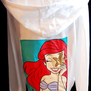 Puede incluir: Sudadera con capucha blanca con un gr&aacute;fico colorido de Ariel de La Sirenita. El gr&aacute;fico muestra a Ariel con cabello rojo, un sujetador de concha p&uacute;rpura y una estrella de mar dorada. La sudadera tiene un fondo turquesa.