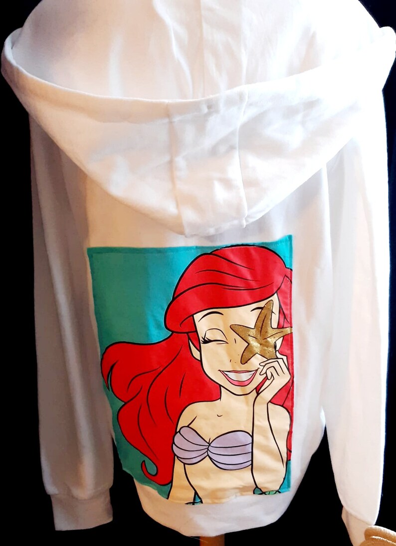 Puede incluir: Sudadera con capucha blanca con un gr&aacute;fico colorido de Ariel de La Sirenita. El gr&aacute;fico muestra a Ariel con cabello rojo, un sujetador de concha p&uacute;rpura y una estrella de mar dorada. La sudadera tiene un fondo turquesa.