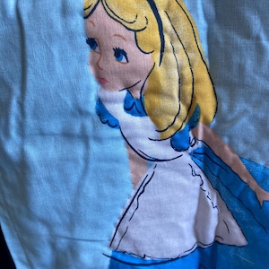 May include: A light blue fabric with an embroidered image of Alice from Alice in Wonderland. Alice has blonde hair, blue eyes, and wears a blue dress with a white apron. A black bow is in her hair.